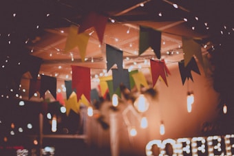 A festive setting adorned with colorful triangular flags hanging from the ceiling. Warm lights twinkle, creating a cozy atmosphere. The blurred background suggests an intimate space, possibly for a celebration or gathering.