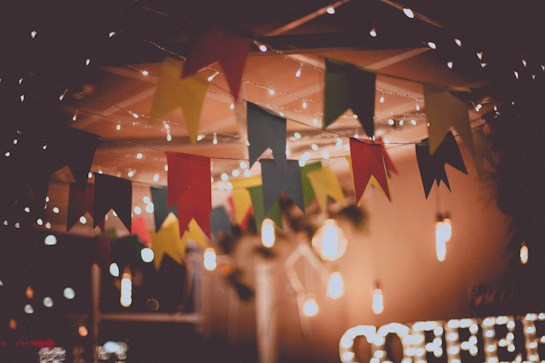 A festive setting adorned with colorful triangular flags hanging from the ceiling. Warm lights twinkle, creating a cozy atmosphere. The blurred background suggests an intimate space, possibly for a celebration or gathering.