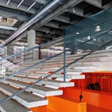 A modern interior space featuring a wide staircase with a concrete and metal design, leading up to an upper level. The stair treads are gray and the risers are orange, contributing to a bold contrasting color scheme. Overhead, there are industrial-style exposed ducts and lighting fixtures. Below the staircase, there is an orange wall with hangers and a wheelchair, and a caution sign with an umbrella is visible.