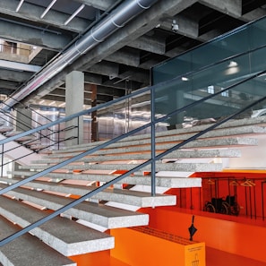 A modern interior space featuring a wide staircase with a concrete and metal design, leading up to an upper level. The stair treads are gray and the risers are orange, contributing to a bold contrasting color scheme. Overhead, there are industrial-style exposed ducts and lighting fixtures. Below the staircase, there is an orange wall with hangers and a wheelchair, and a caution sign with an umbrella is visible.
