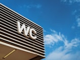 An outdoor sign with the letters 'WC' is mounted on a modern structure with horizontal slats, set against a clear blue sky with wispy clouds.