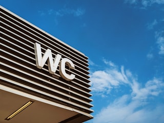 An outdoor sign with the letters 'WC' is mounted on a modern structure with horizontal slats, set against a clear blue sky with wispy clouds.
