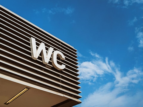 An outdoor sign with the letters 'WC' is mounted on a modern structure with horizontal slats, set against a clear blue sky with wispy clouds.