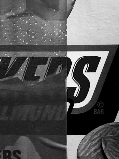 A close-up view of packaging with large, bold text partially visible. The design features a mix of patterns, with dots and textured surfaces. The dominant color scheme is grayscale.