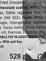 A close-up view of printed text on packaging, providing ingredient information for a filled chocolate product. The text includes details about chocolate coating, edible vegetable fats, sugar, and allergen warnings. There is visible wear and tear on the packaging, indicating possible crumpling or damage.