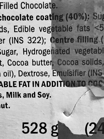 A close-up view of printed text on packaging, providing ingredient information for a filled chocolate product. The text includes details about chocolate coating, edible vegetable fats, sugar, and allergen warnings. There is visible wear and tear on the packaging, indicating possible crumpling or damage.