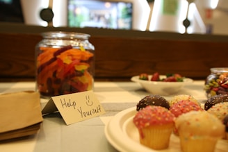 A table is set with a variety of sweets, including a jar of colorful gummy candies, a plate with small cupcakes topped with different colored icing and sprinkles, and a bowl of mixed berries. A handwritten note on the table says 'Help Yourself' with a smiley face.