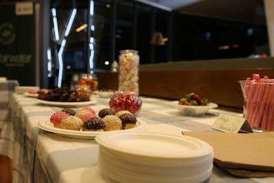 A variety of sweets and treats are displayed on a checkered tablecloth. There are cupcakes with colorful sprinkles, a bowl of strawberries, assorted cookies, and a jar of marshmallows. Pink straws are arranged in a holder, and there is a sign that reads 'Help Yourself.' The setting is indoors with a dimly lit background.