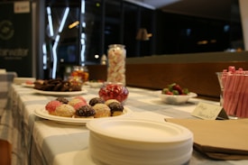 A variety of sweets and treats are displayed on a checkered tablecloth. There are cupcakes with colorful sprinkles, a bowl of strawberries, assorted cookies, and a jar of marshmallows. Pink straws are arranged in a holder, and there is a sign that reads 'Help Yourself.' The setting is indoors with a dimly lit background.