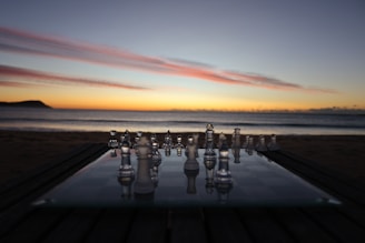 A glass chessboard is set up on a table outdoors at sunset, with the ocean and a colorful sky in the background. The chess pieces are reflective, capturing the hues of the sunset.