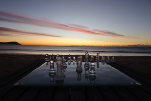 A glass chessboard is set up on a table outdoors at sunset, with the ocean and a colorful sky in the background. The chess pieces are reflective, capturing the hues of the sunset.