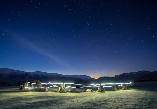 A serene, nighttime landscape featuring a stone circle in an open field. A clear night sky filled with stars serves as the backdrop, with distant hills visible on the horizon. A trail of light weaves through the stone circle, suggesting movement, possibly created by a long exposure photograph.