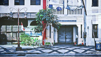An urban street scene features a vintage building with weathered textures and a large metal door marked with the year 1918. A bicycle with a basket is parked nearby, alongside a small tree and Indonesian flag. The area is adorned with a classic street lamp and various urban elements like a trash bin and cables.