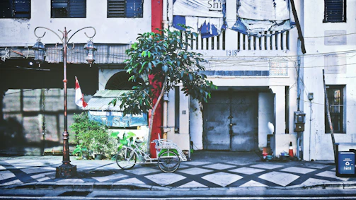 An urban street scene features a vintage building with weathered textures and a large metal door marked with the year 1918. A bicycle with a basket is parked nearby, alongside a small tree and Indonesian flag. The area is adorned with a classic street lamp and various urban elements like a trash bin and cables.