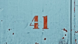 The image depicts the number 41 stenciled in red on a worn, weathered metal surface with a faded blue paint background. The surface appears to be aged, with visible rust spots and minor scratches dispersed across the metal.