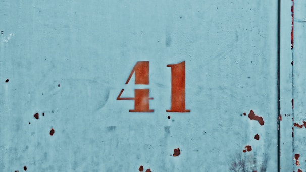 The image depicts the number 41 stenciled in red on a worn, weathered metal surface with a faded blue paint background. The surface appears to be aged, with visible rust spots and minor scratches dispersed across the metal.