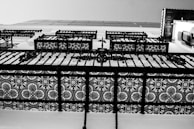 A close-up view of an intricately designed iron balcony railing featuring symmetric floral patterns. The image is in black and white, emphasizing the contrast between the ornate details and the background.