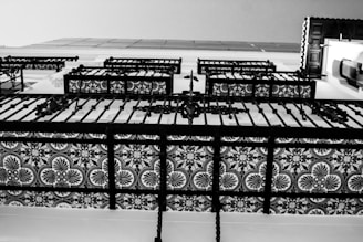 A close-up view of an intricately designed iron balcony railing featuring symmetric floral patterns. The image is in black and white, emphasizing the contrast between the ornate details and the background.