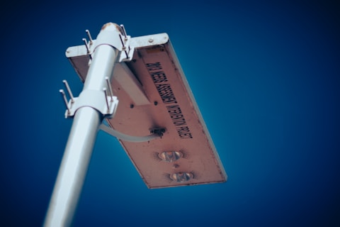 A metal pole extends upwards, holding a rectangular solar panel against a clear, bright blue sky. The panel has an inscription that reads 'PM3 WEED ASSESSMENT VIBRATION PROJECT' and features several metallic components bolted onto its underside.