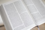 An open book with closely printed text in small font on its pages, showing a passage from the Gospel according to Mark. The book is open to pages with two columns of text, and there is a ribbon bookmark visible at the bottom.