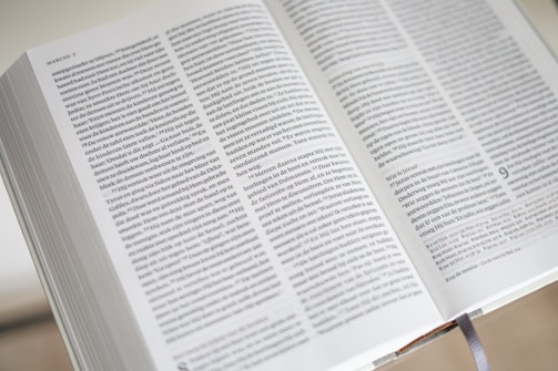 An open book with closely printed text in small font on its pages, showing a passage from the Gospel according to Mark. The book is open to pages with two columns of text, and there is a ribbon bookmark visible at the bottom.