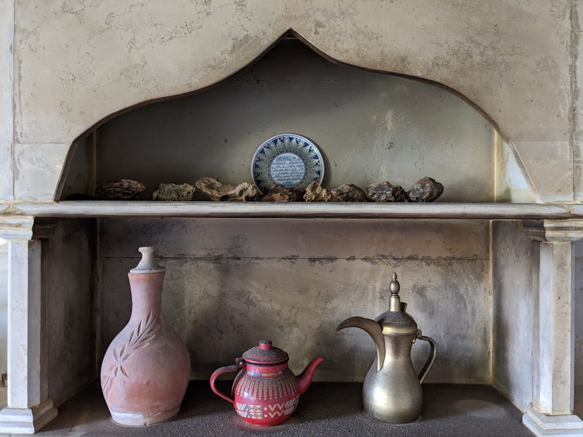 A niche in a stone wall features a decorative arrangement of objects. The upper shelf displays a ceramic plate with intricate blue and white designs, surrounded by a collection of small rocks. The lower shelf holds three vessels: a tall clay jug with leaf imprints, a red teapot with geometric white patterns, and a metallic pitcher with a long spout and bulbous top.