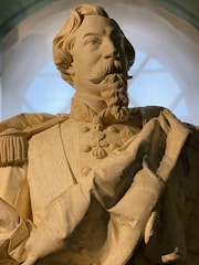A detailed marble statue of a man with a mustache and goatee, wearing a decorated military uniform with epaulettes and a large medallion on the chest. The figure has an intricate hairstyle and is portrayed looking upwards with a contemplative expression. The background shows a softly lit arch or window.