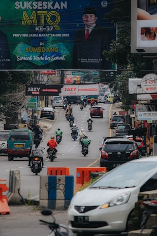 A busy urban street featuring several motorcyclists and vehicles moving in both directions. Above the road, numerous billboards are displayed, including a prominent sign advertising an event in Sumedang. The atmosphere is lively with the hustle and bustle typical of a city setting.
