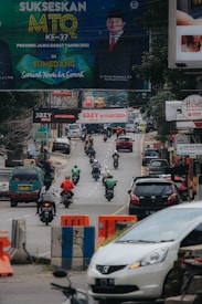 A busy urban street featuring several motorcyclists and vehicles moving in both directions. Above the road, numerous billboards are displayed, including a prominent sign advertising an event in Sumedang. The atmosphere is lively with the hustle and bustle typical of a city setting.