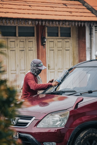 A person wearing a hat and face covering is cleaning the windshield of a red Honda car parked in front of a building with an orange tiled roof and cream-colored doors.