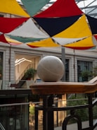 A large, smooth, spherical stone is placed on a wooden railing inside a modern building. Overhead, colorful triangular fabric panels in red, yellow, black, and green are suspended, allowing light to pass through. The background includes stairs, lush plants, and glass storefronts, enhancing the architectural appeal.