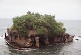 A small, rocky island with lush green foliage and a traditional, pagoda-like structure at the top. The island is surrounded by the ocean, and the waves gently crash against its sides. Stone steps and pathways are visible around the edges, leading to the natural formations and structures on the island.