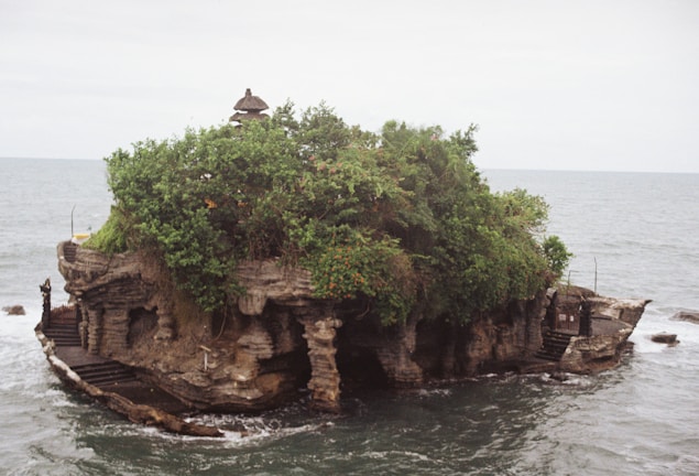 A small, rocky island with lush green foliage and a traditional, pagoda-like structure at the top. The island is surrounded by the ocean, and the waves gently crash against its sides. Stone steps and pathways are visible around the edges, leading to the natural formations and structures on the island.