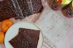 A white plate with a piece of dark brown food, possibly jerky or dried meat, is placed on a light surface. Surrounding the plate are several packets of similar dark brown food items, some packaged in transparent plastic. There are also two bright orange tangerines on the left side of the plate. To the right, a package with a branded wrapper labeled 'FYRE' and a few jars with colorful designs feature prominently.