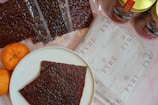 A white plate with a piece of dark brown food, possibly jerky or dried meat, is placed on a light surface. Surrounding the plate are several packets of similar dark brown food items, some packaged in transparent plastic. There are also two bright orange tangerines on the left side of the plate. To the right, a package with a branded wrapper labeled 'FYRE' and a few jars with colorful designs feature prominently.