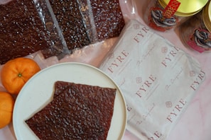 A white plate with a piece of dark brown food, possibly jerky or dried meat, is placed on a light surface. Surrounding the plate are several packets of similar dark brown food items, some packaged in transparent plastic. There are also two bright orange tangerines on the left side of the plate. To the right, a package with a branded wrapper labeled 'FYRE' and a few jars with colorful designs feature prominently.