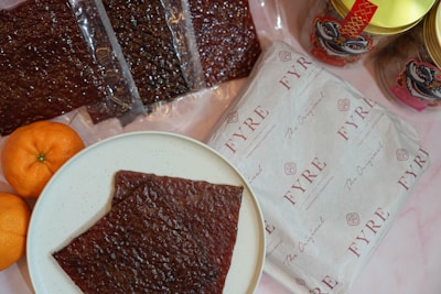 A white plate with a piece of dark brown food, possibly jerky or dried meat, is placed on a light surface. Surrounding the plate are several packets of similar dark brown food items, some packaged in transparent plastic. There are also two bright orange tangerines on the left side of the plate. To the right, a package with a branded wrapper labeled 'FYRE' and a few jars with colorful designs feature prominently.