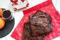 Several pieces of dark, grilled meat are laid out on red paper with a traditional pattern. To the side, there is a glass filled with red wine on a black slate coaster, accompanied by two oranges. A decorative bakery box with an elaborate design is partially visible in the background.