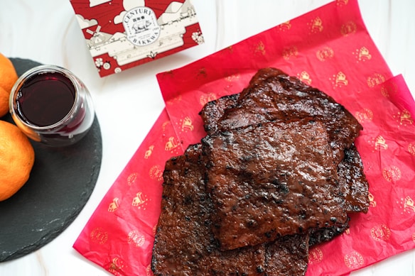 Several pieces of dark, grilled meat are laid out on red paper with a traditional pattern. To the side, there is a glass filled with red wine on a black slate coaster, accompanied by two oranges. A decorative bakery box with an elaborate design is partially visible in the background.