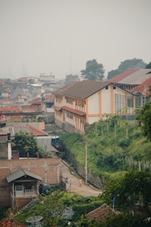 A suburban or rural neighborhood with a mix of modern and traditional architecture. A long building with a brown roof and light yellow walls stands prominently, surrounded by greenery and smaller houses with red-tiled roofs. A narrow road winds through the area, where a person in a yellow shirt is seen walking. The sky is overcast, creating a muted and serene atmosphere.