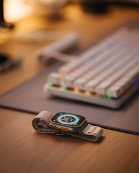 A smartwatch with a digital display showing various metrics is resting on a desk beside a mechanical keyboard with colorful keycaps. The wooden surface and soft ambient lighting create a cozy setting.