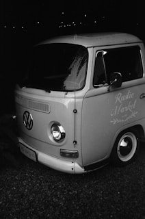 A black-and-white image of a vintage Volkswagen van with the words 'Rustic Market' on the side. The van is parked on a gravel surface, and its classic design features round headlights and a split windshield.