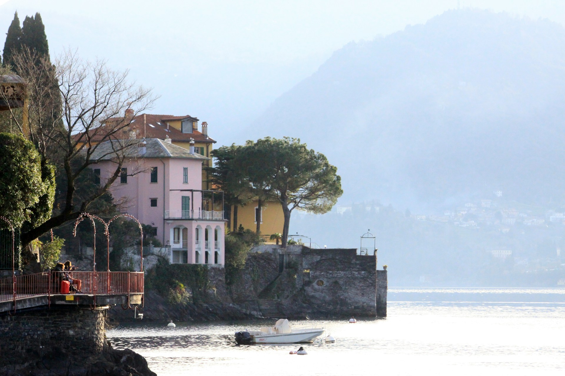A serene lakeside view with elegant villas perched on the shore, surrounded by lush greenery. The calm water reflects the soft light of the sky as a small boat rests near the edge. The backdrop features misty, rolling hills that add depth to the scene. Leafless trees and manicured gardens frame the buildings, giving a touch of elegance and tranquility.