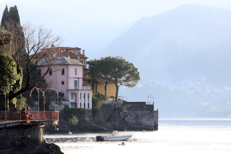 A serene lakeside view with elegant villas perched on the shore, surrounded by lush greenery. The calm water reflects the soft light of the sky as a small boat rests near the edge. The backdrop features misty, rolling hills that add depth to the scene. Leafless trees and manicured gardens frame the buildings, giving a touch of elegance and tranquility.