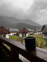 A black mug is placed on a wooden railing, with a background of rustic wooden cabins and a misty mountain landscape. The atmosphere is cloudy and overcast, suggesting rain or impending rain.
