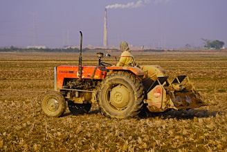 A person wearing headgear operates an orange tractor in a vast, plowed agricultural field. A tall smokestack emits smoke in the background, with a hazy sky and some distant buildings and power lines visible. The soil looks dry and the landscape is expansive.