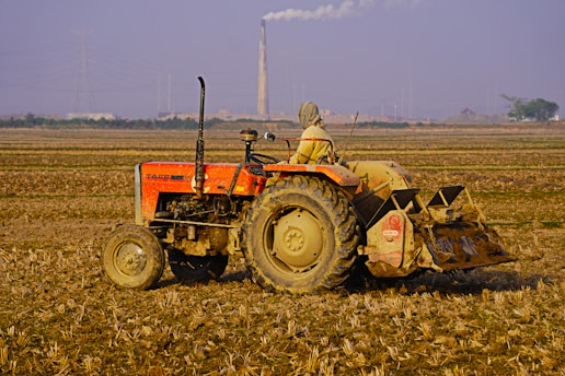 A person wearing headgear operates an orange tractor in a vast, plowed agricultural field. A tall smokestack emits smoke in the background, with a hazy sky and some distant buildings and power lines visible. The soil looks dry and the landscape is expansive.