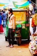A man wearing casual clothing and sandals stands next to a green auto rickshaw labeled CNG in a busy marketplace. Multiple people are visible in the background, along with various colorful stalls and canopies. The scene appears vibrant and bustling with activity.