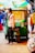 A man wearing casual clothing and sandals stands next to a green auto rickshaw labeled CNG in a busy marketplace. Multiple people are visible in the background, along with various colorful stalls and canopies. The scene appears vibrant and bustling with activity.