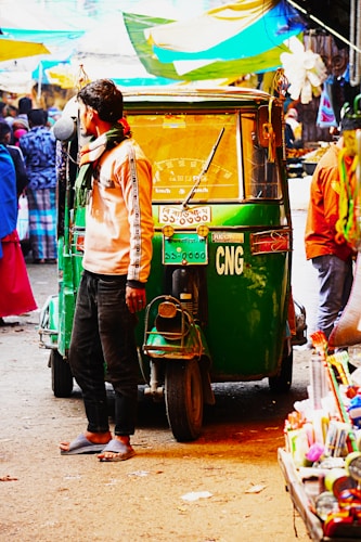 A man wearing casual clothing and sandals stands next to a green auto rickshaw labeled CNG in a busy marketplace. Multiple people are visible in the background, along with various colorful stalls and canopies. The scene appears vibrant and bustling with activity.